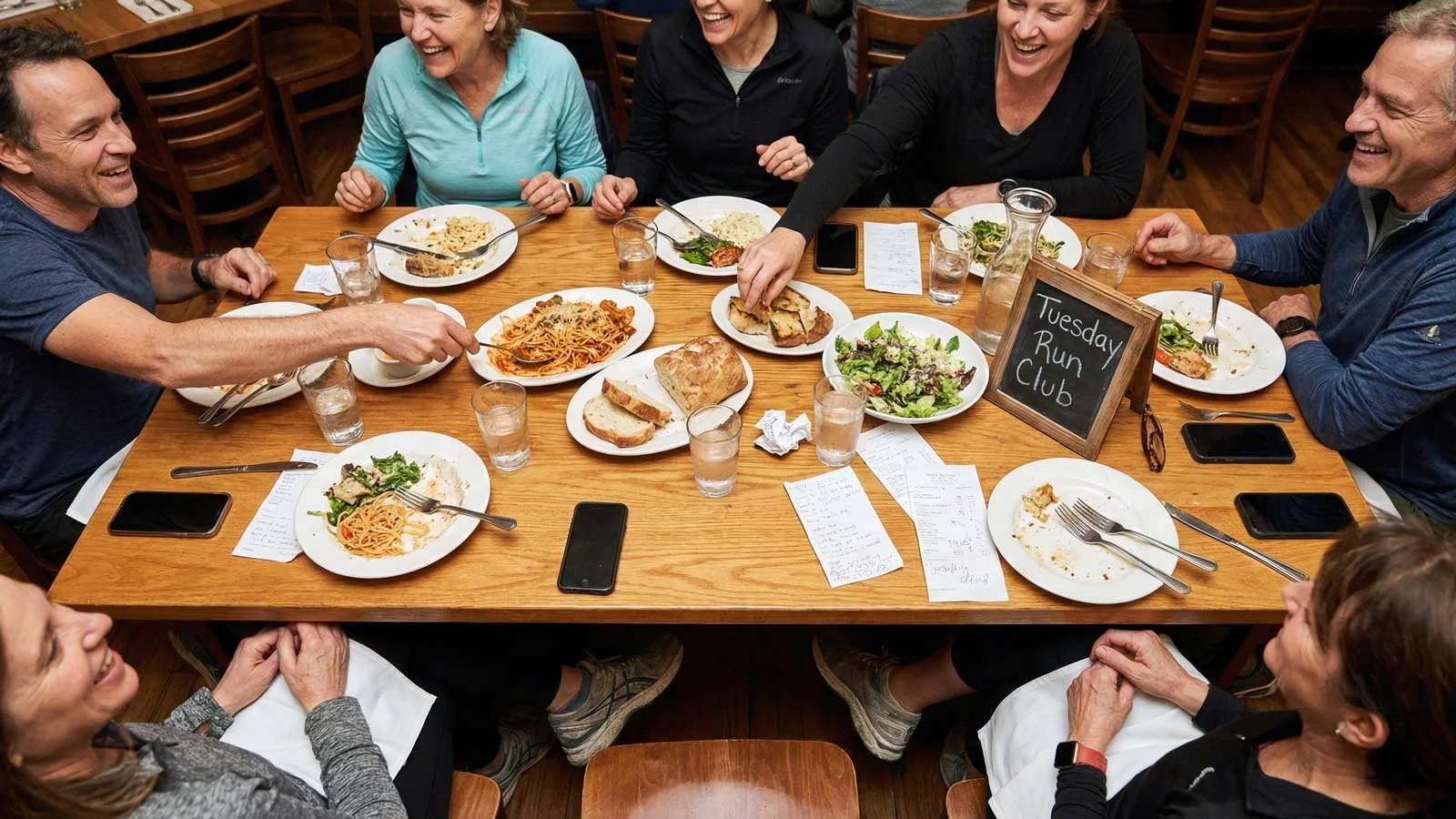 Overhead shot of a crowded neighbourhood restaurant table with running shoes tucked under chairs, phones face-down on the table, hands reaching for shared plates, handwritten notes on receipts, and a small chalkboard sign reading 'Tuesday Run Club'.